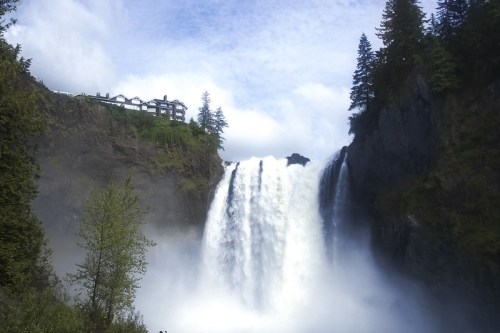 A powerful waterfall cascades down rocky cliffs, surrounded by lush greenery and a distant building atop the hill.