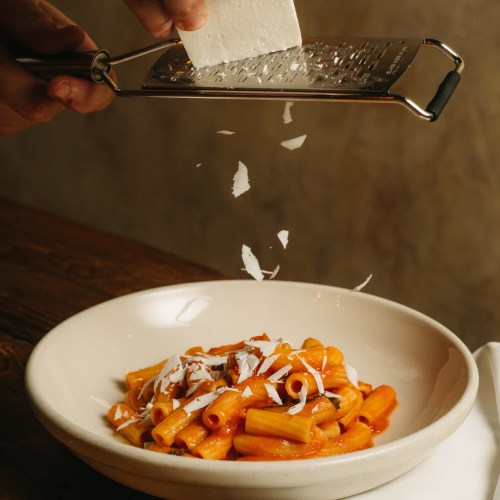 A hand grating cheese over a bowl of pasta with tomato sauce on a wooden table.