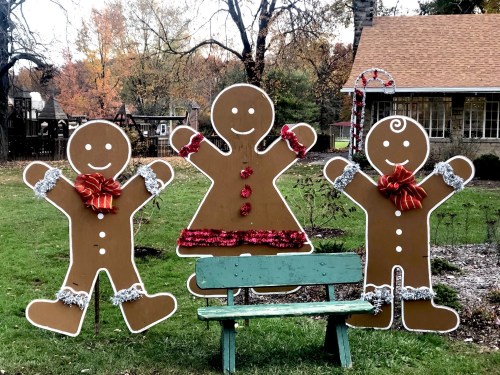 Three large gingerbread figures, two with red bows and one in a dress, stand beside a green bench in a festive outdoor setting.