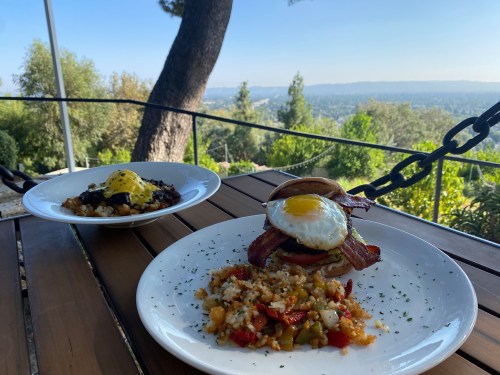 Two plates of breakfast on a wooden table, featuring a sunny-side-up egg, bacon, and a view of greenery and hills.