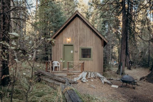A small wooden cabin in a forest, with a green door, a chair on the porch, and a fire pit nearby.