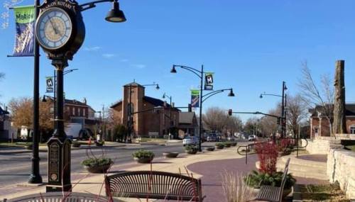A street view featuring a vintage clock, benches, and banners in a small town with buildings in the background.