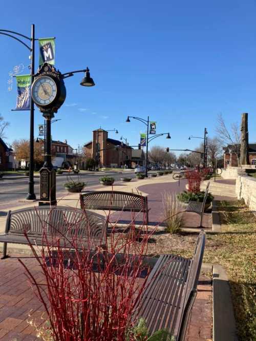 A sunny street scene featuring benches, decorative plants, and a clock, with banners hanging along the road.