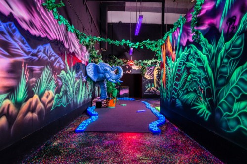 A vibrant, neon-lit hallway with jungle-themed murals, an elephant statue, and colorful, sparkly flooring.