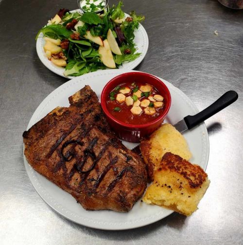 A plate with a grilled steak, cornbread, a bowl of chili, and a side salad with apples and nuts.