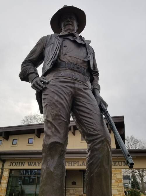 A large statue of a cowboy holding a rifle, in front of the John Wayne Birthplace Museum.