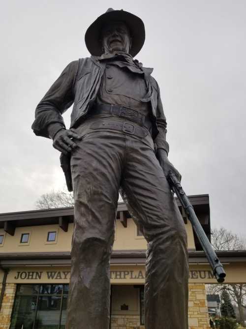 A large statue of a cowboy holding a rifle, in front of the John Wayne Birthplace Museum.