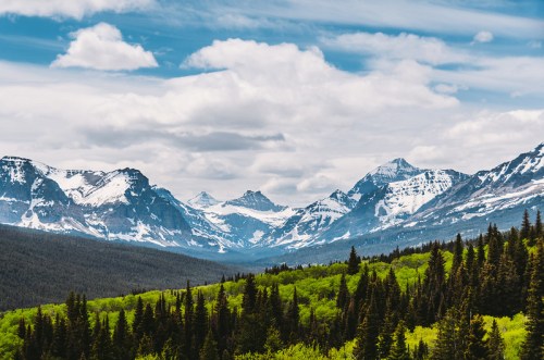 A scenic view of snow-capped mountains under a blue sky, surrounded by lush green forests.