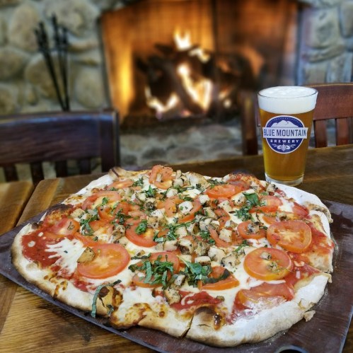 A freshly baked pizza topped with tomatoes and herbs, next to a pint of beer, with a cozy fireplace in the background.