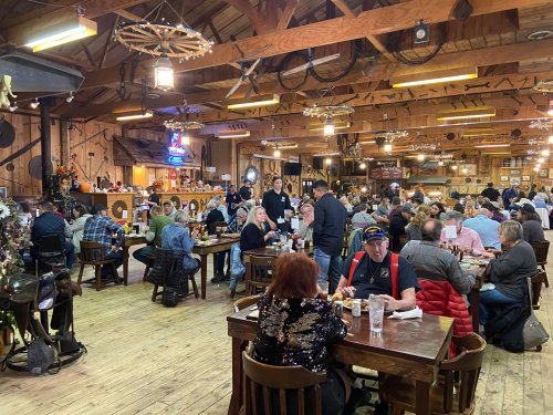 A bustling restaurant interior with wooden decor, diners at tables, and staff serving food in a lively atmosphere.