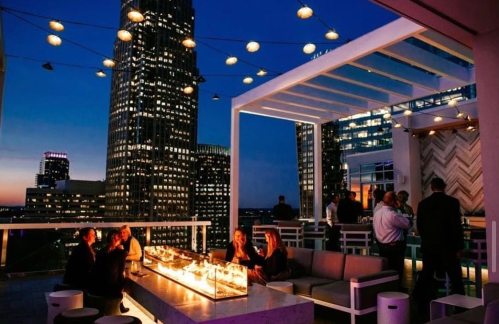 A rooftop lounge at dusk, featuring a fire pit, seating, and city skyline views with twinkling lights.