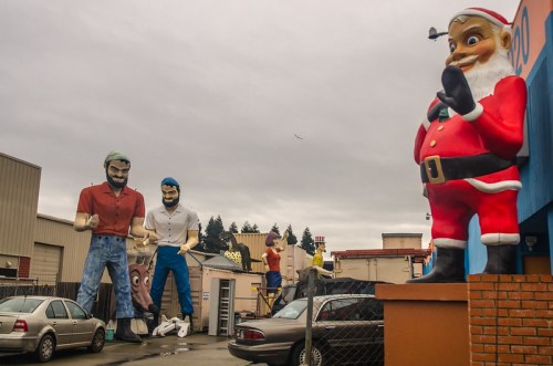 A collection of large, whimsical statues, including Santa Claus and two fishermen, in an industrial area under a cloudy sky.