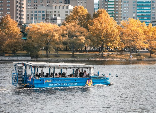 A blue amphibious tour boat navigates the water with autumn trees and city buildings in the background.