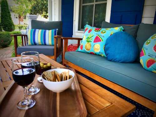 A cozy outdoor seating area with colorful cushions, two glasses of red wine, and a bowl of snacks on a wooden tray.