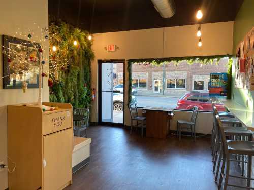 A cozy café interior with greenery, a thank you sign, and seating by a large window overlooking the street.