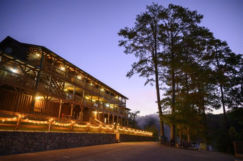 A rustic lodge illuminated by string lights, surrounded by tall trees at dusk.