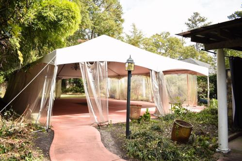 A large white tent with clear sides set in a garden, surrounded by greenery and a pathway leading to it.