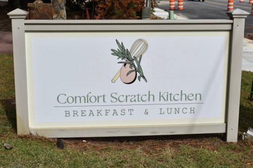 Sign for "Comfort Scratch Kitchen" featuring a whisk and herbs, indicating breakfast and lunch offerings.