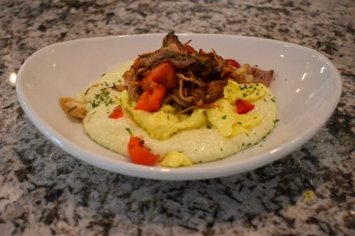 A bowl of creamy grits topped with pulled meat, diced tomatoes, and herbs on a marble countertop.