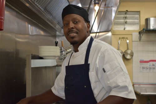 A chef in a kitchen, wearing a white uniform and black hat, smiles while preparing food at a stainless steel counter.