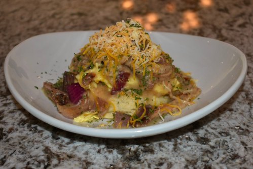 A plate of creamy mashed potatoes topped with cheese, sautéed vegetables, and herbs, served in a white bowl.