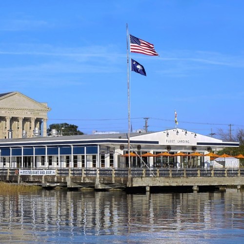 A waterfront restaurant with an American flag and a South Carolina flag, surrounded by calm water and blue skies.