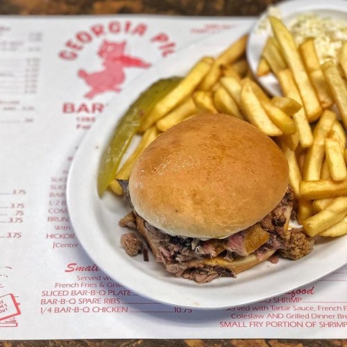 A plate featuring a pulled pork sandwich, fries, pickles, and a menu backdrop from Georgia Pig BBQ.