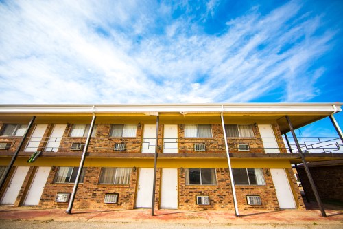 A low-angle view of a brick motel with multiple rooms and a blue sky filled with clouds.