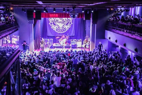 A lively concert scene with a large crowd enjoying a band on stage, illuminated by colorful lights.