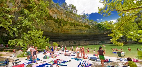 A sunny beach scene with people swimming and relaxing by a green river, surrounded by cliffs and lush trees.
