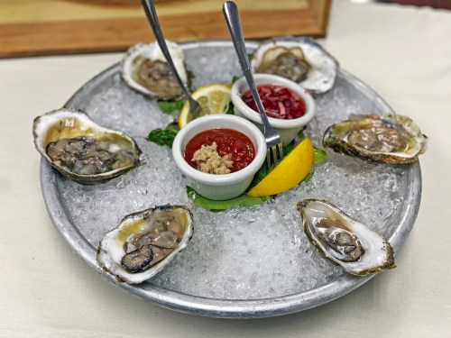 A platter of fresh oysters on ice, served with lemon wedges and dipping sauces in small bowls.