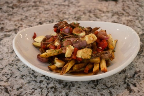 A bowl of poutine featuring fries topped with beef, cheese curds, tomatoes, and onions on a marble countertop.
