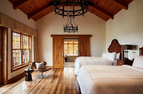 A cozy bedroom with two beds, wooden beams, large windows, and rustic decor, featuring a chair and side table.