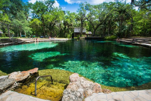 A serene natural spring surrounded by lush greenery and clear blue water, with people enjoying the area.