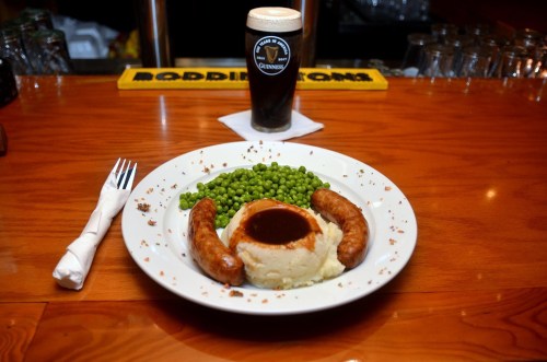 A plate with sausages, mashed potatoes with gravy, and green peas, alongside a pint of Guinness beer.