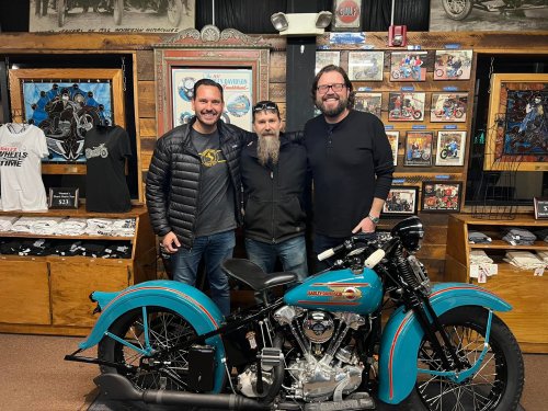 Three men pose together in a motorcycle shop, standing beside a vintage turquoise motorcycle.
