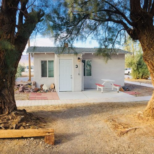 A small white house with a door marked "3," framed by two large trees and a gravel pathway.
