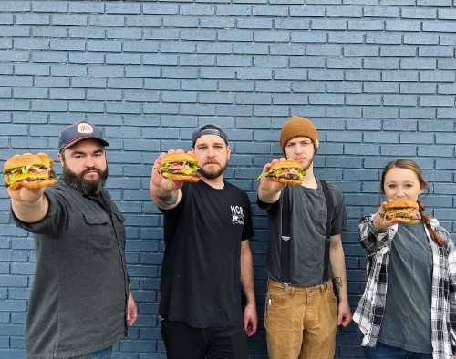 Four people stand in front of a blue brick wall, each holding a burger and smiling at the camera.