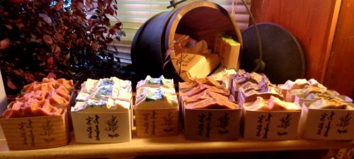A display of colorful handmade soaps arranged neatly on a wooden shelf, with a rustic barrel in the background.