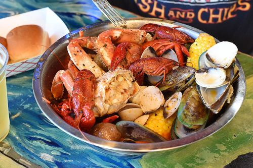 A metal bowl filled with various seafood, including lobster, crawfish, clams, mussels, and corn, on a colorful table.