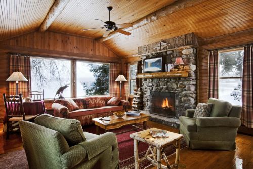 Cozy cabin living room with a stone fireplace, wooden ceiling, and large windows overlooking a snowy landscape.