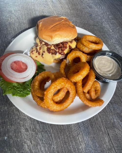 A plate with a cheeseburger topped with bacon, onion rings, lettuce, tomato, and a side of dipping sauce.