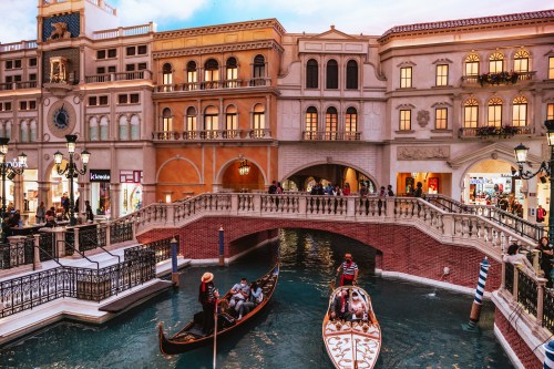 A scenic view of a Venetian-style canal with gondolas, bridges, and shoppers in a vibrant indoor shopping area.