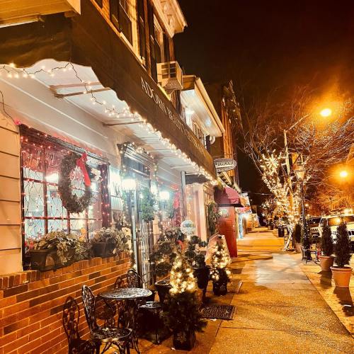 A charming street scene at night, adorned with festive lights and decorations, featuring small tables and trees.