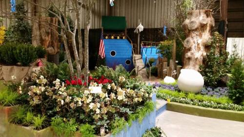 A vibrant garden scene featuring colorful flowers, greenery, and a blue boat structure with an American flag.