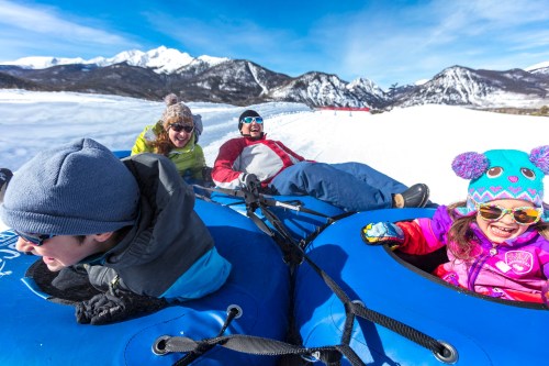 A family enjoys snow tubing on a sunny day, surrounded by snowy mountains and blue skies. Kids and adults are smiling.