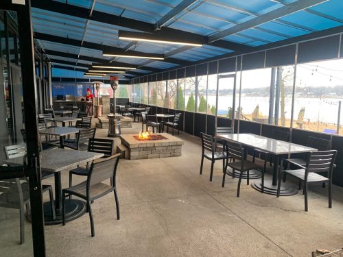 A covered outdoor dining area with tables, chairs, and a fire pit, overlooking a lake.