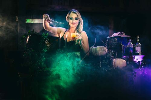 A woman in a dark dress holds a glowing drink, surrounded by smoke and autumn decorations, creating a mystical atmosphere.