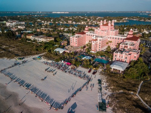 Aerial view of a pink hotel by the beach, with a volleyball court and people gathered on the sandy shore.