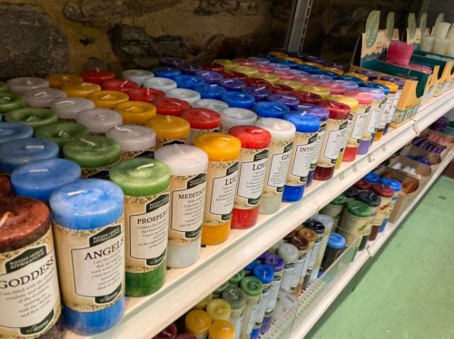 A colorful display of various candles on shelves, labeled with names like "Goddess," "Angels," and "Prosperity."